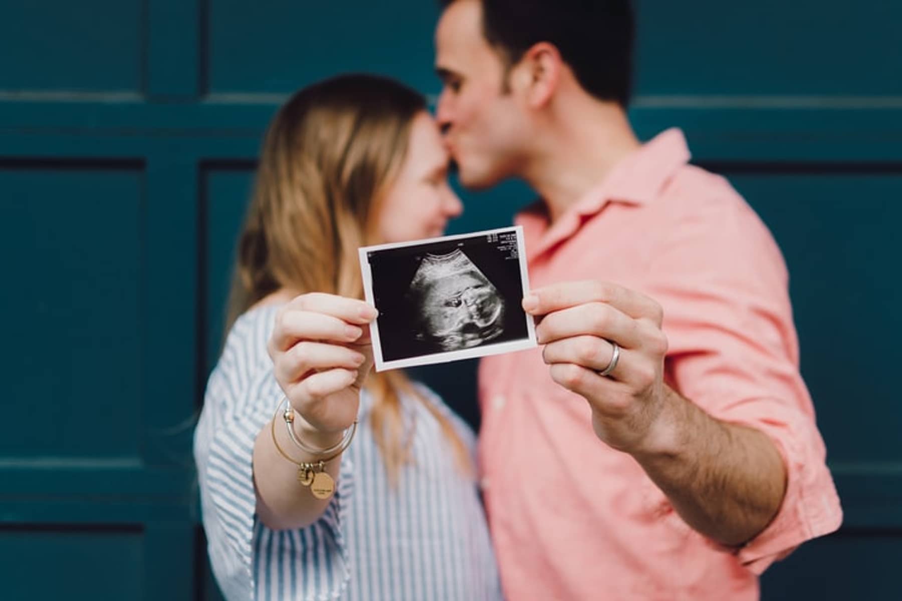 A couple holding an ultrasound image, with the man kissing the woman's forehead, in front of a blue door.