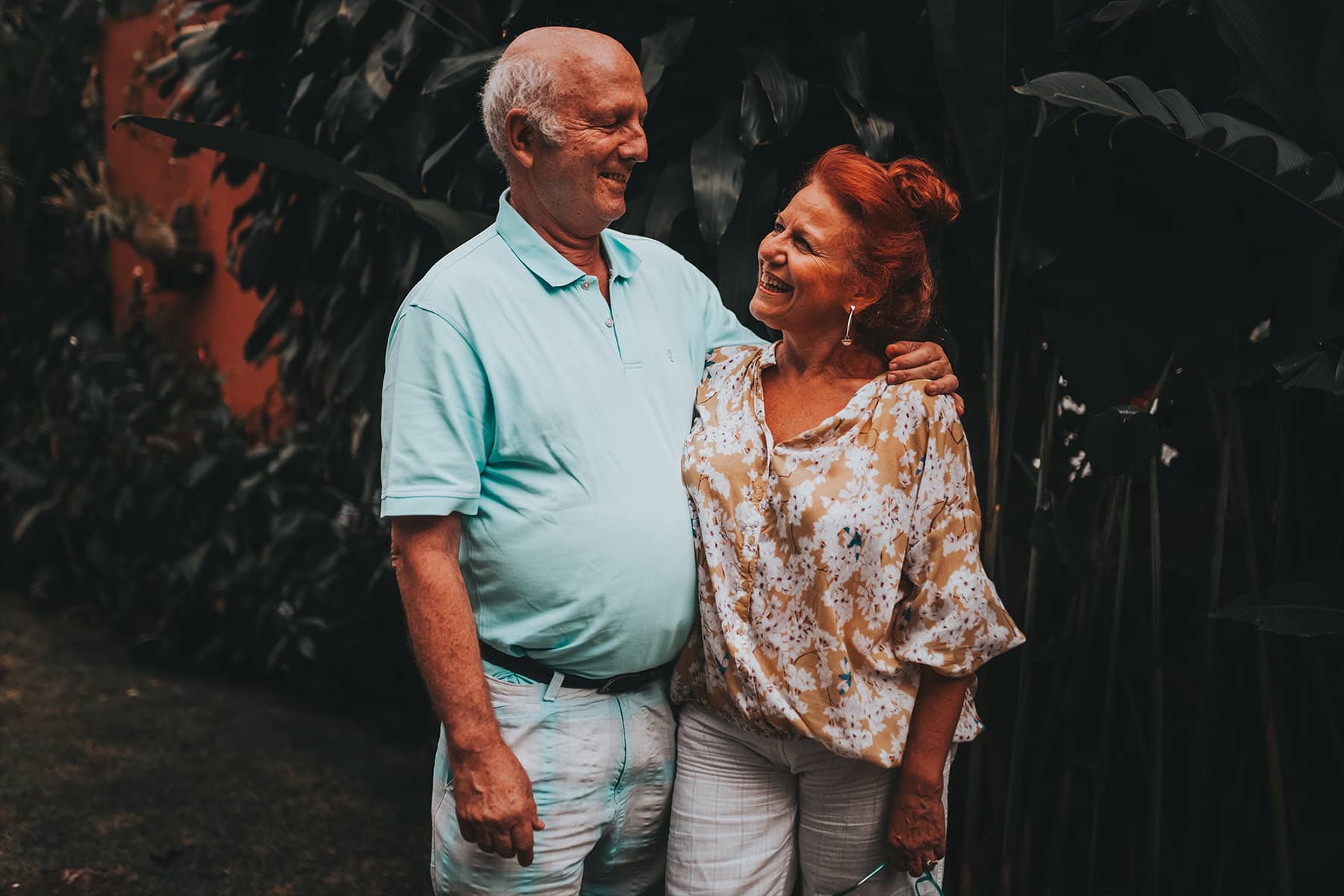 An older couple smiling at each other, with the man's arm around the woman, set against a backdrop of dark greenery.