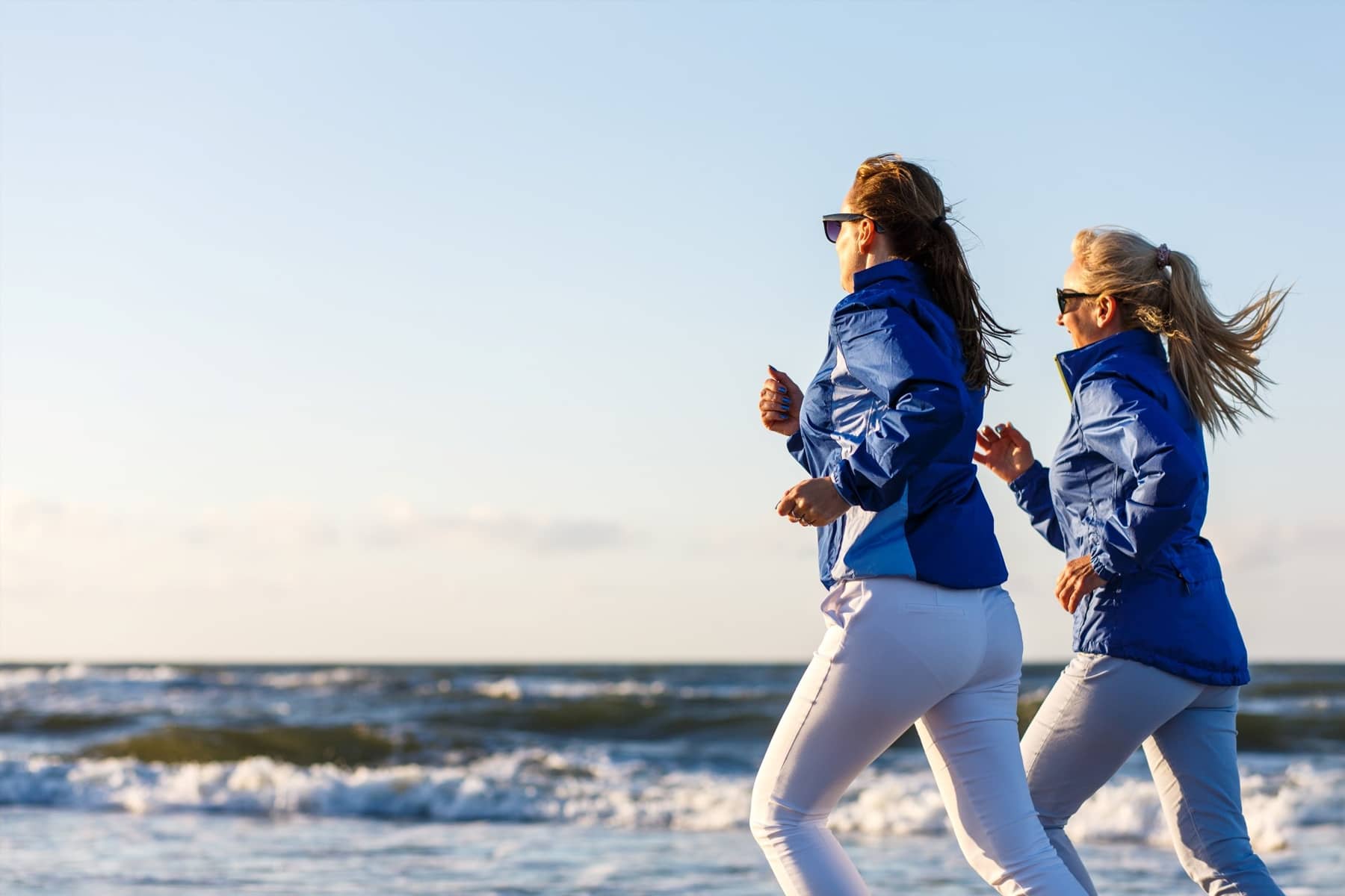 Two women jogging along a beach, wearing blue jackets and white pants, with waves and the sky in the background.