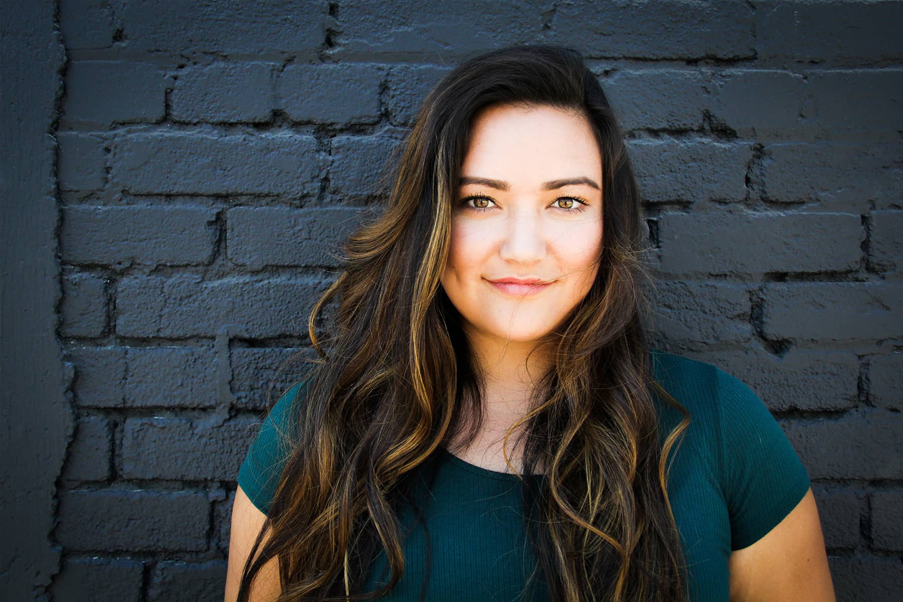 Headshot of a woman with long, wavy brown hair and blonde highlights, wearing a green top, against a dark gray brick wall.