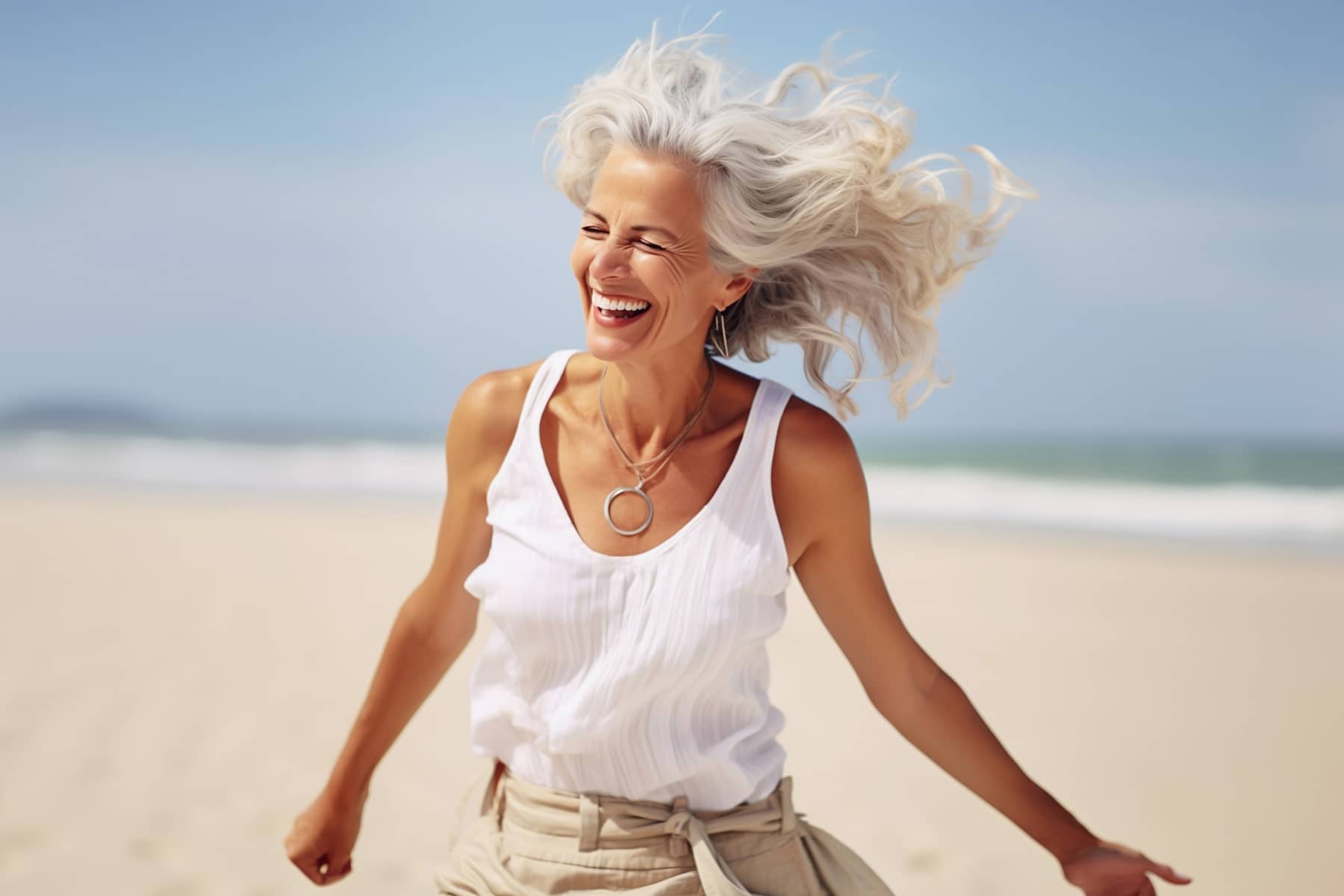 A mature woman with gray hair laughing joyfully on a beach, with her hair blowing in the wind.