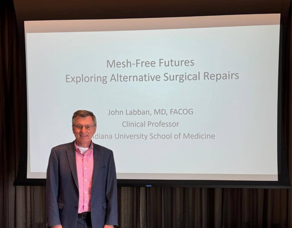 Dr. John Labban stands confidently in front of a large screen displaying the title "Mesh-Free Futures: Exploring Alternative Surgical Repairs" and his credentials.