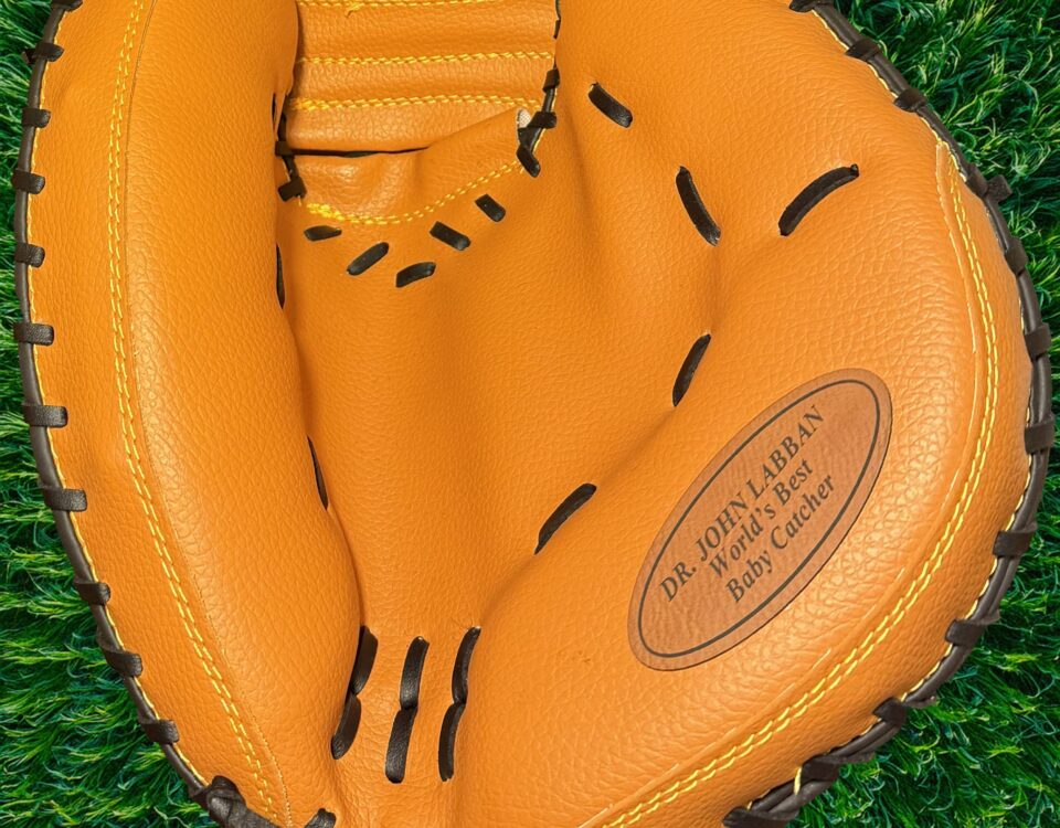 Close-up of a tan leather baseball catcher's mitt engraved with “Dr. John Labban – World’s Best Baby Catcher,” resting on green artificial turf.