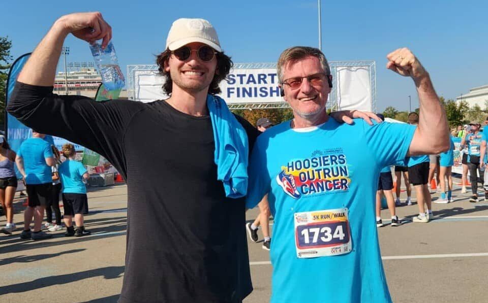 Two men smiling and flexing their arms after finishing the Hoosiers Outrun Cancer 5K event, surrounded by other participants near the finish line.